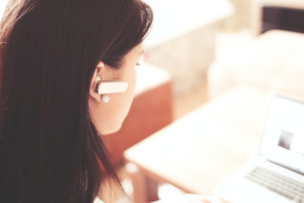 Woman wearing earpiece using a white laptop computer.