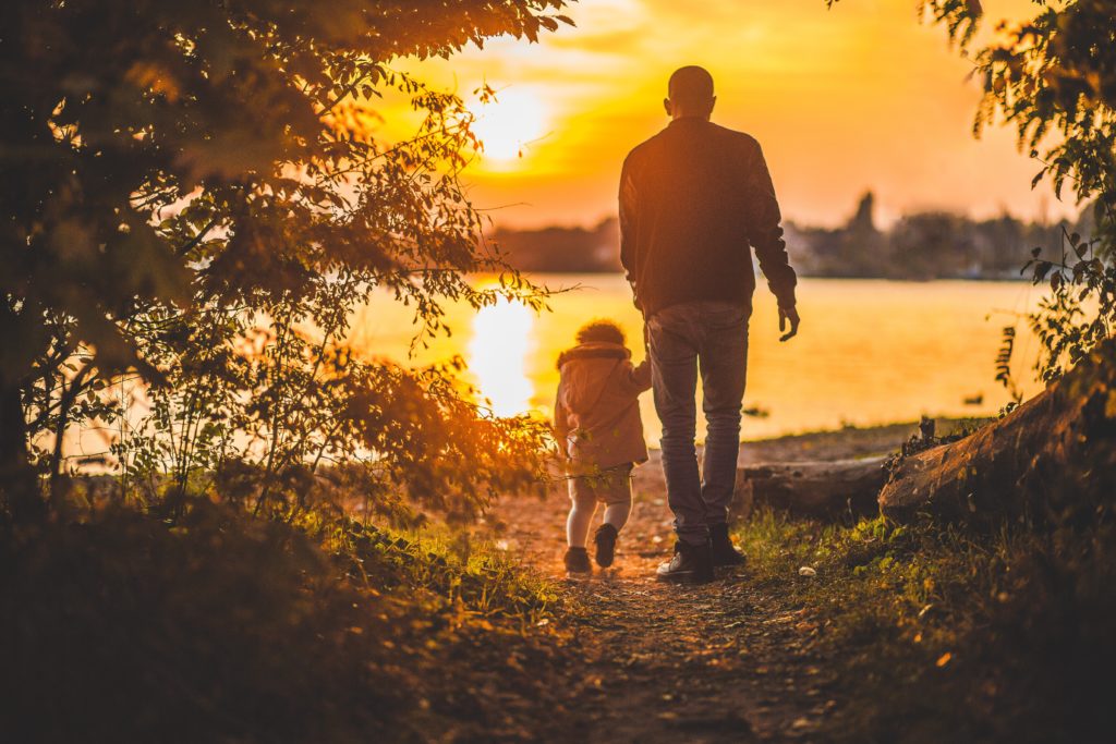 A father and daughter walking hand in hand toward a sunset over a lake.
