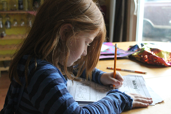 A young girl with a blue striped long-sleeved shirt completing an activity sheet at a dinner table in front of a doorwall.