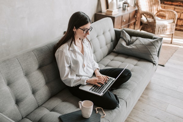 Woman wearing white shirt working on a laptop while sitting on a grey couch