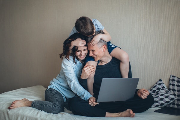 A mother and father sitting on a bed with their son laughing while on a laptop.