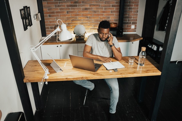 A man working on a laptop while talking on the phone on a wood desk.
