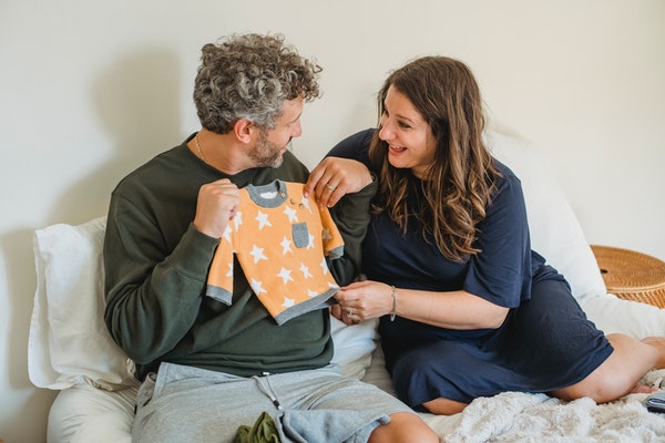 Husband and wife sitting on bed holding up baby clothes