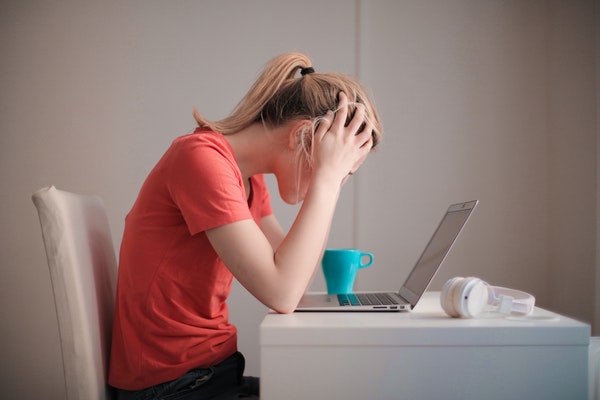 Girl wearing coral shirt leaned over a laptop with hands on her head in despair