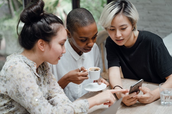 Three women sitting at a cafe table drinking espresso and looking at a cellphone.