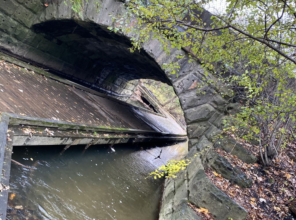 A wood plank walkway along a creek under a stone bridge.