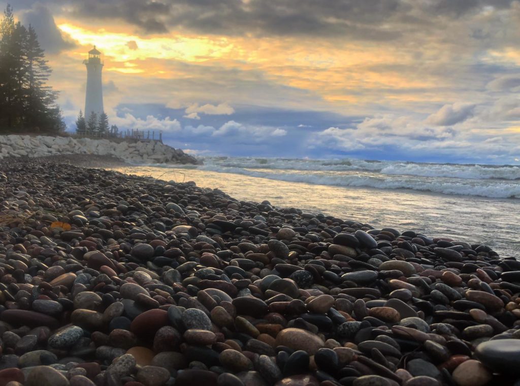 Waves crashing on a rocky shore in Newberry, Michigan with a lighthouse in the background.