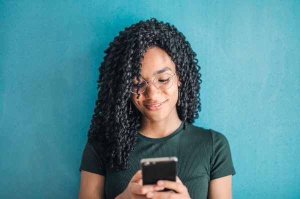 African American female with glasses leaning against teal wall looking at her phone.