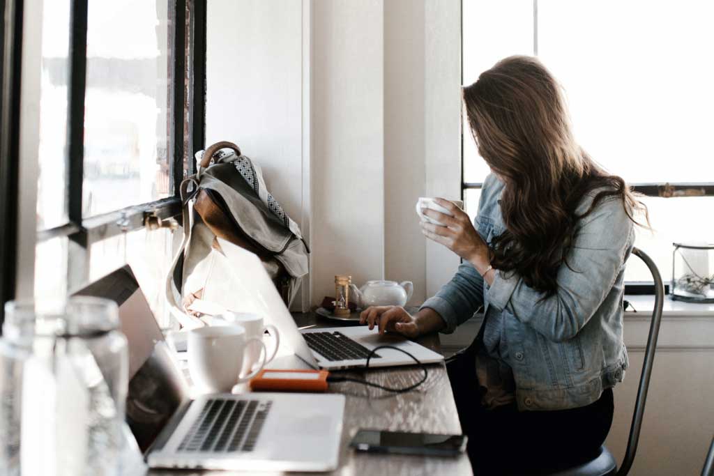 A woman holding a cup of coffee while she works from a computer in a coffee shop.
