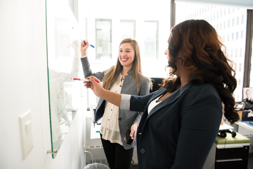 Two female professionals brainstorming on a whiteboard in an office.