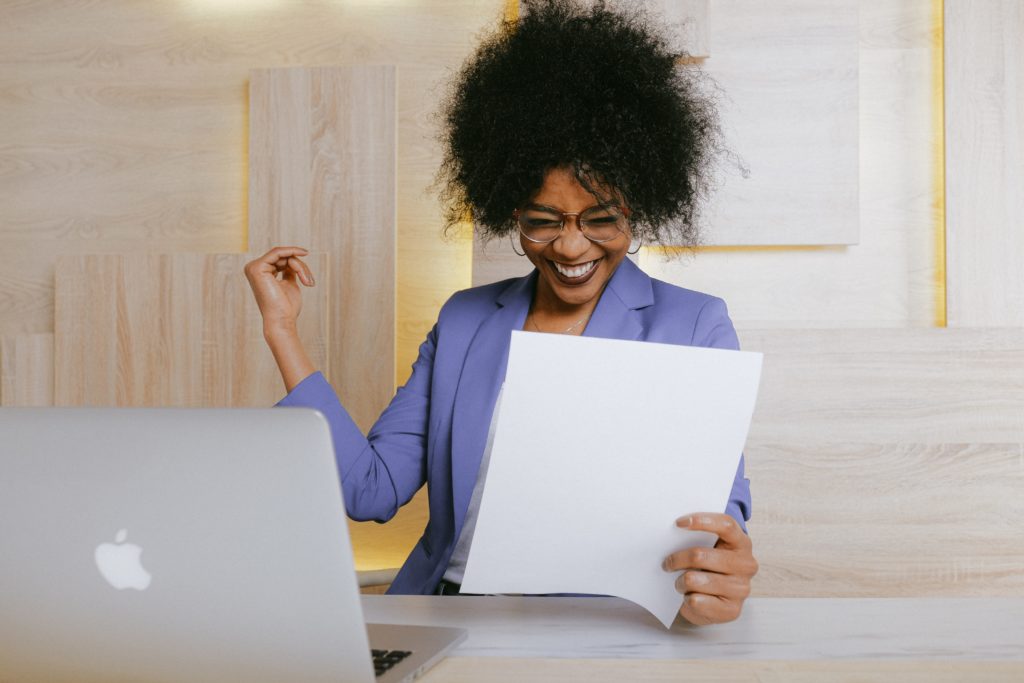 Woman sitting at a desk holding a piece of paper and excitedly smiling.