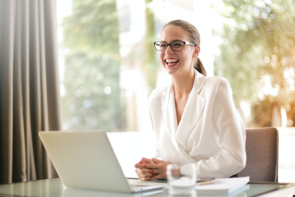 Smiling blonde woman sitting at a desk with a computer as she greets a guest.
