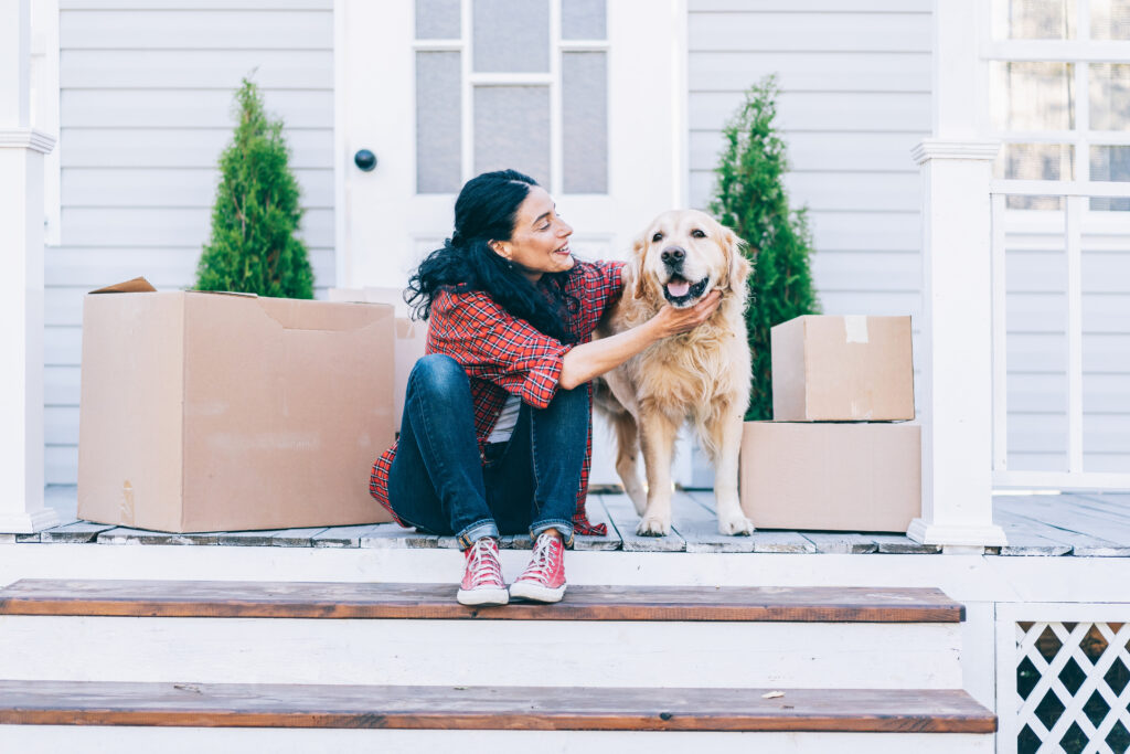Woman sitting on a front porch with a dog surrounded by cardboard boxes.