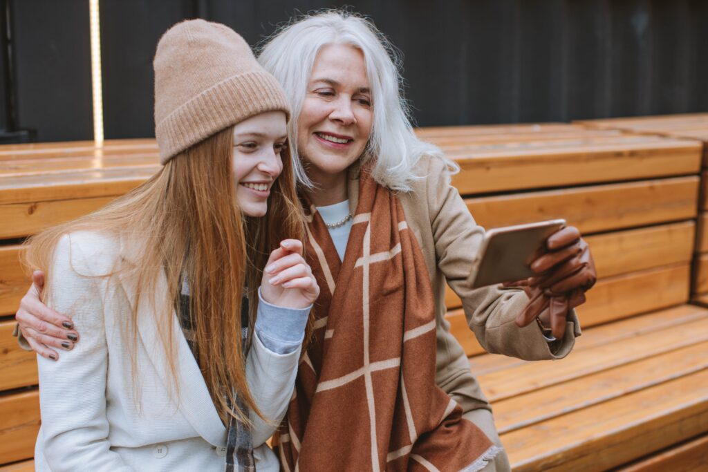 An older woman and younger woman smile for the camera as they take a selfie on a park bench.