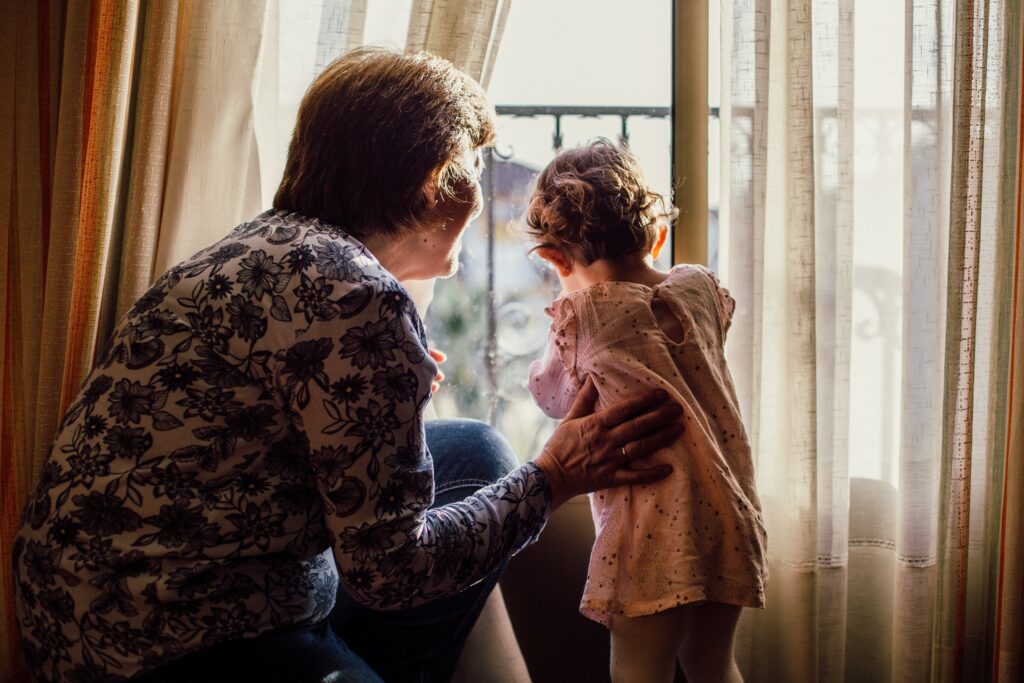 An elderly woman helps a young child as they peer out a window.