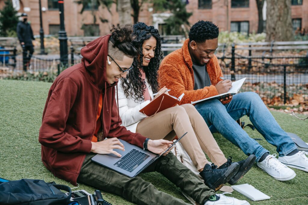 Three college students, two male and one female, sit on a hill as they study from open text books.