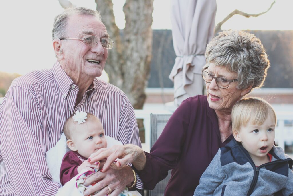 An older couple hold their young great-grandchildren as they sit outside.