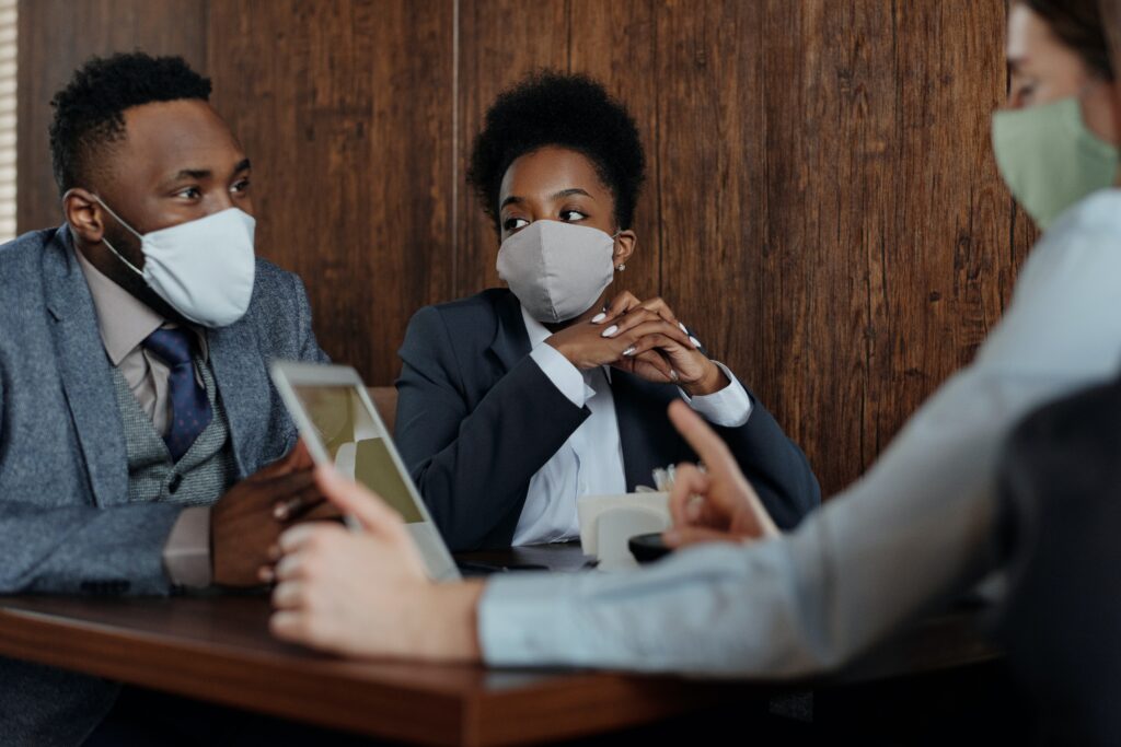 Three people in professional business attire and masks seated around a table having a discussion.