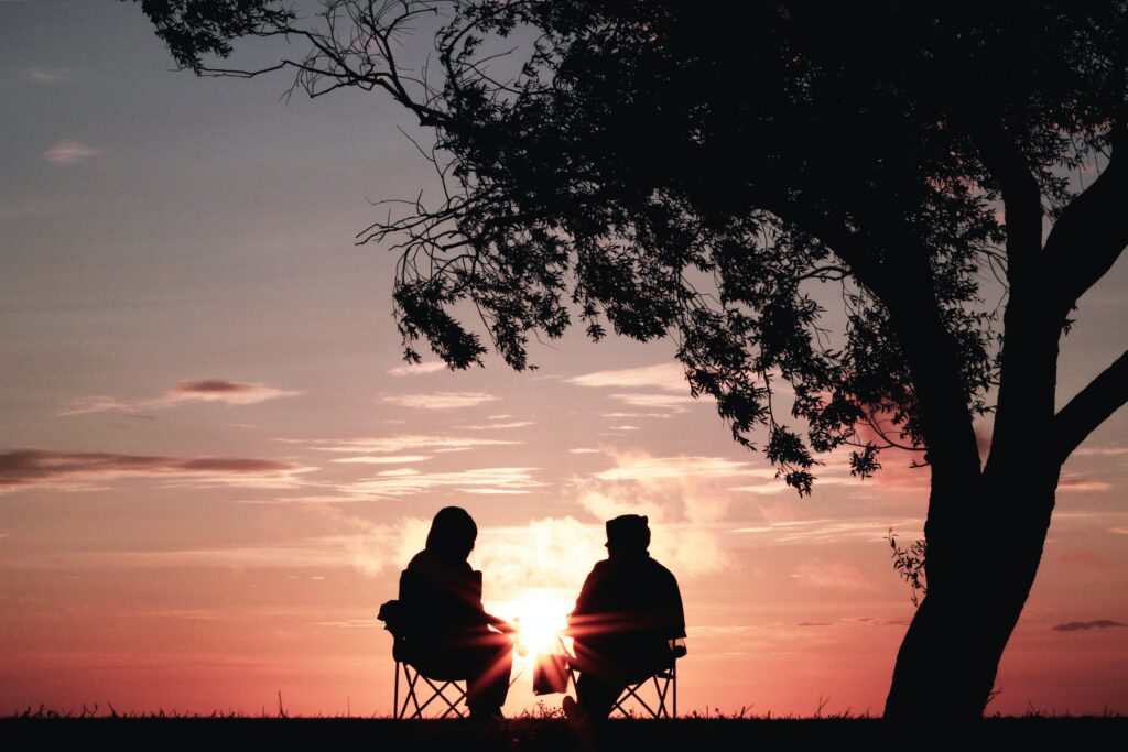 Two people sit in lawn chairs, under a tree as they admire a sunset sky.
