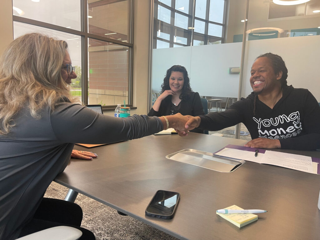 Dondrea Brown and Lynne Jarman-Johnson shake hands across a table as they solidify their financial education partnership while Stephanie Stacey smiles int he background.