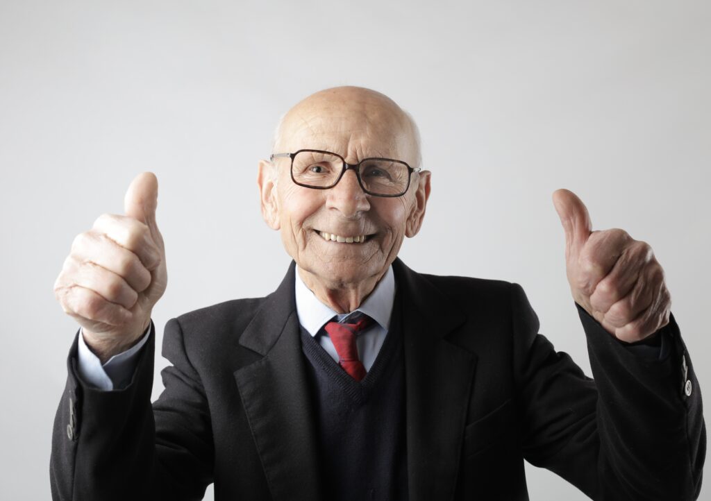 An older man in a business suit smiling widely as he holds up two thrumbs-up.