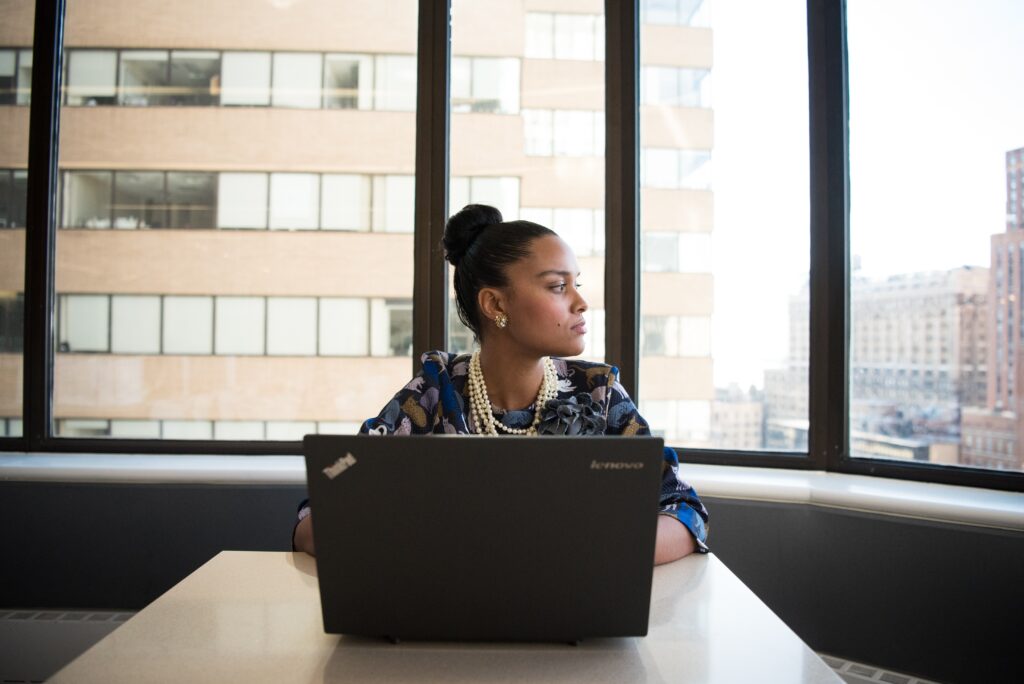 A business woman sitting at a conference desk in front of an open computer as she looks out the window.