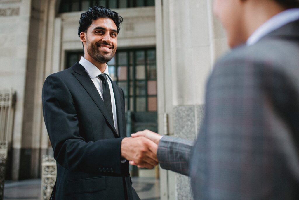 A man wearing a suit shakes hands with another business professional in front of a large building.