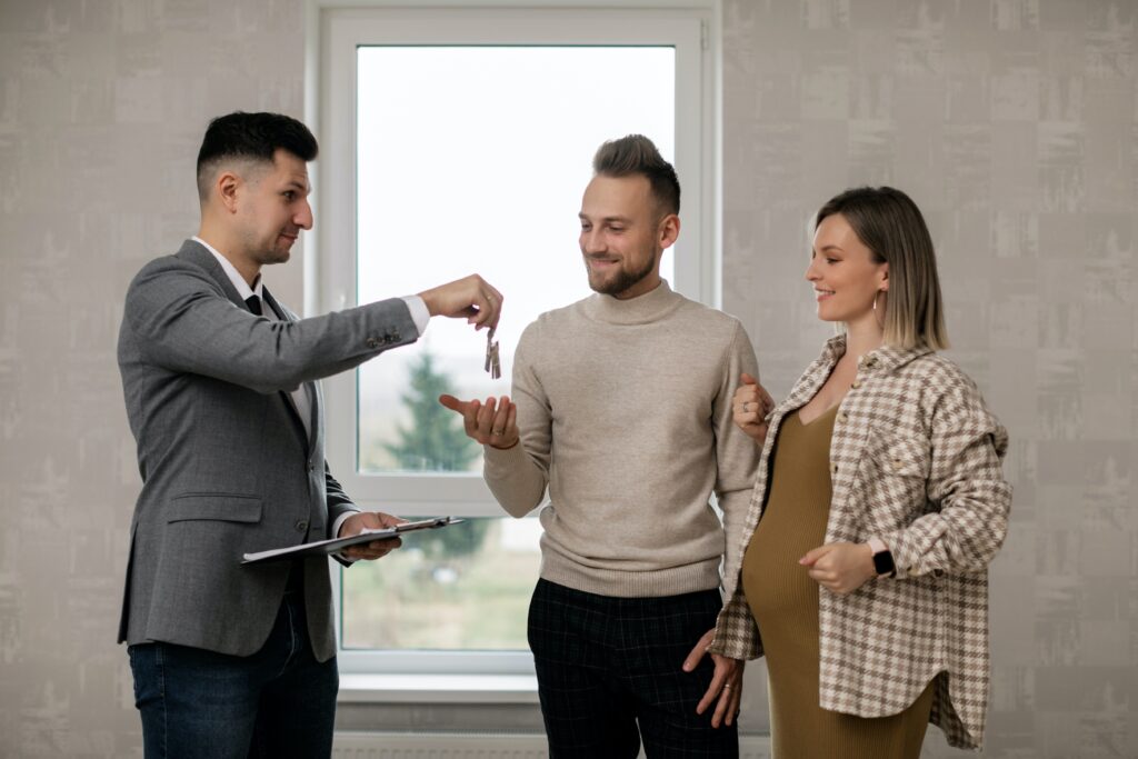 A male realtor handing a set of house keys over to a smiling couple expecting a baby.
