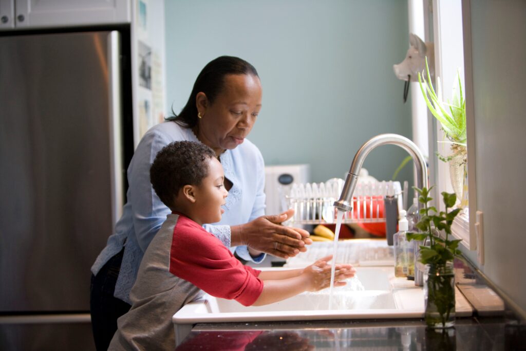 An adult woman helping a young child wash his hands in the kitchen sink.