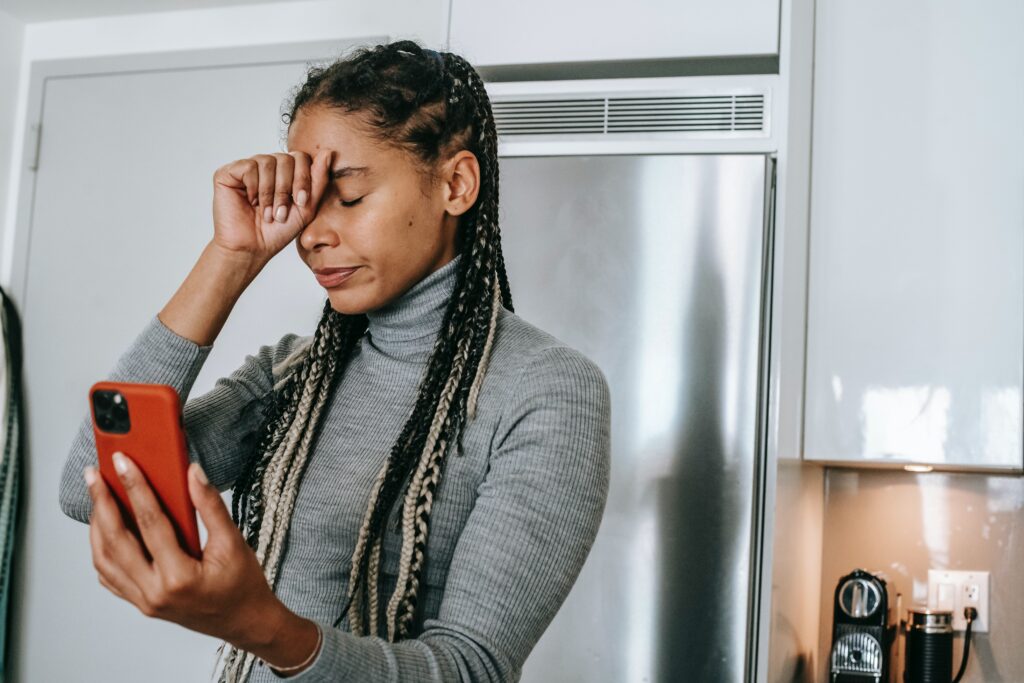 A woman standing in her kitchen holding a red phone as she looks at the screen and puts her hand on her head in frustration.