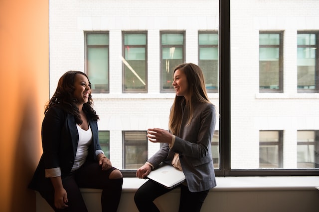 Two female business professionals sit across from each other in a window sill as they collaborate on a project.