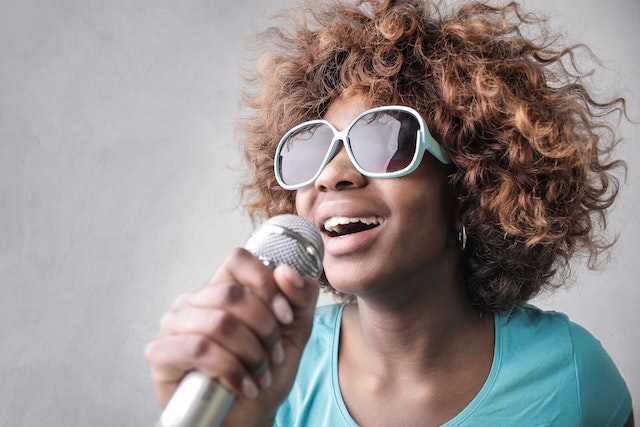 A woman wearing sunglasses smiles as she sings into a microphone.