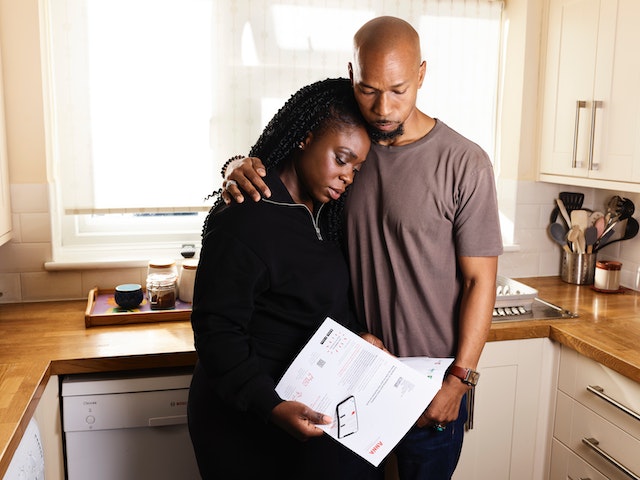 A couple embraces as they comfort each other while standing in a kitchen holding a piece paper.