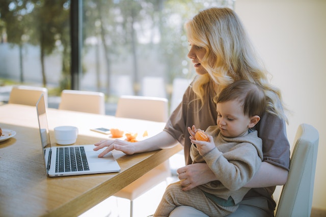 A woman with blonde hair sitting at a table as she works from her open computer and holds a baby on her lap.