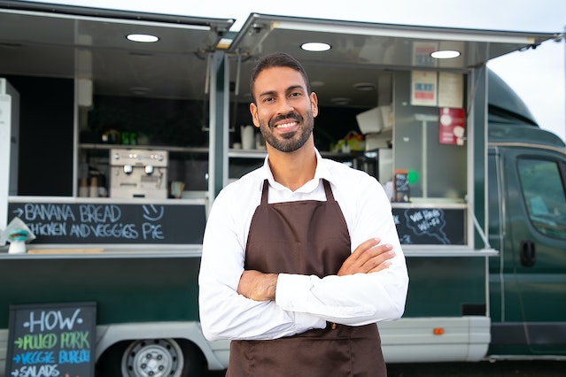 A man wearing an apron smiles in front of his open food truck business.