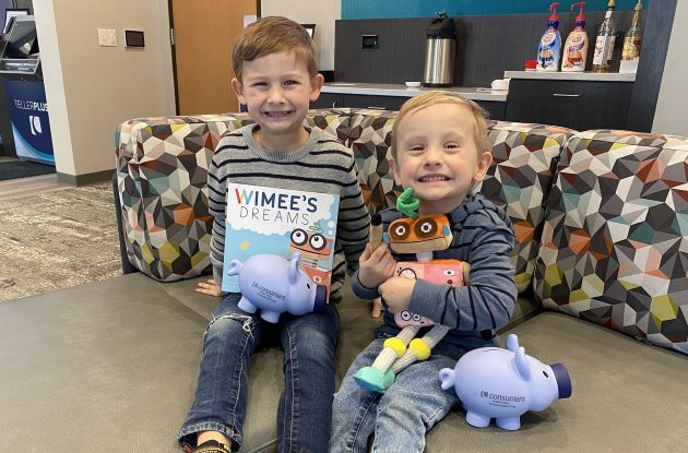 Two young boys sitting on a couch holding a Wimee book and plush