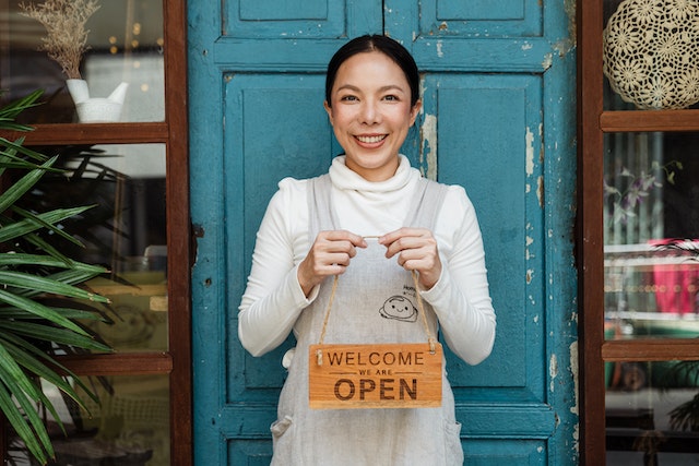 A business owner stands in front of the blue door to her business as she holds a sign that reads "Welcome! We're open!"