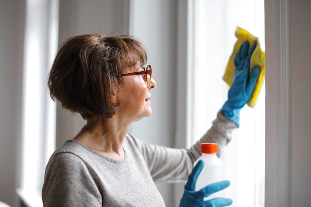 A middle-aged woman with short, brown hair and eyeglasses is cleaning a window with a cloth and cleaning solution.
