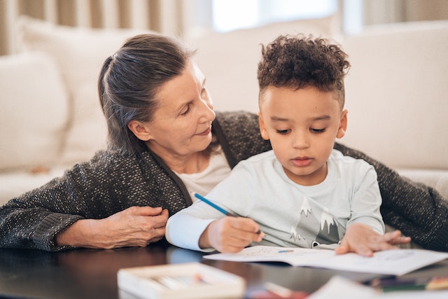 A middle-aged woman sits next to her young grandson as he colors on paper.