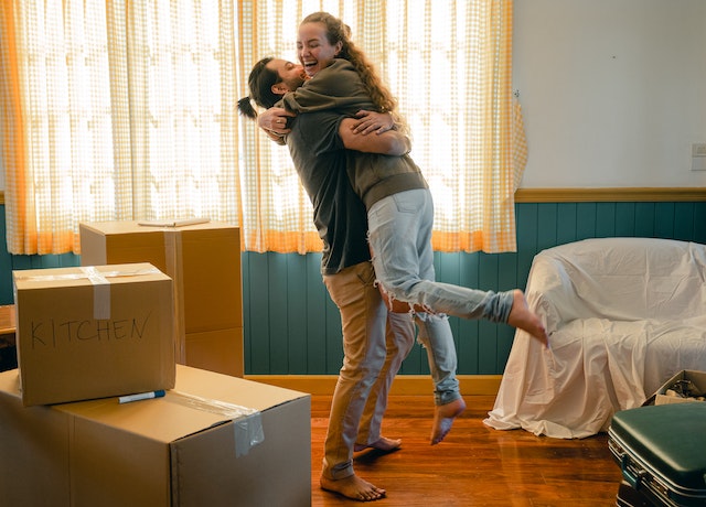 A male and female couple happily embrace in a room filled filled with moving boxes.