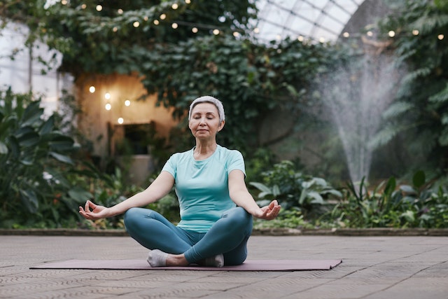 An older woman meditating in a garden as she sits on a yoga mat.