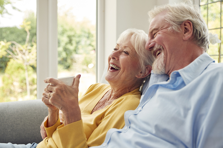 An elderly couple sit near each other on a couch as they drink coffee.