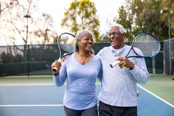 An elderly couple embrace on a tennis court while the rest rackets over their shoulders.