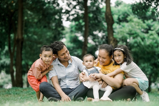 A family of five sit together on the green grass as they smile at the camera.