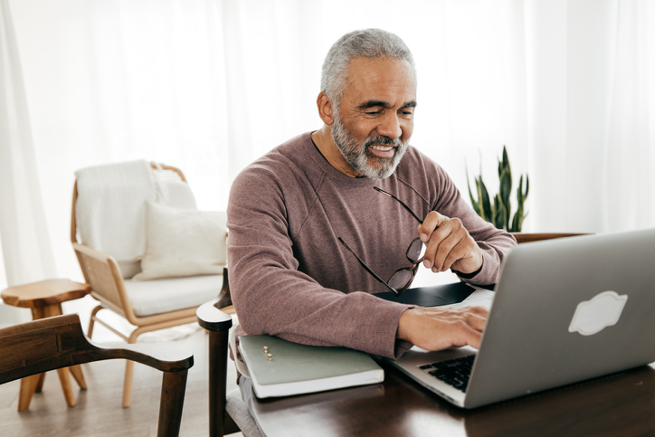 A middle-aged man with graying hair smiles down at the screen of his computer while holding a pair of eyeglasses.