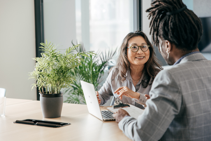 Two business professionals chat at a conference table in front of an open computer.