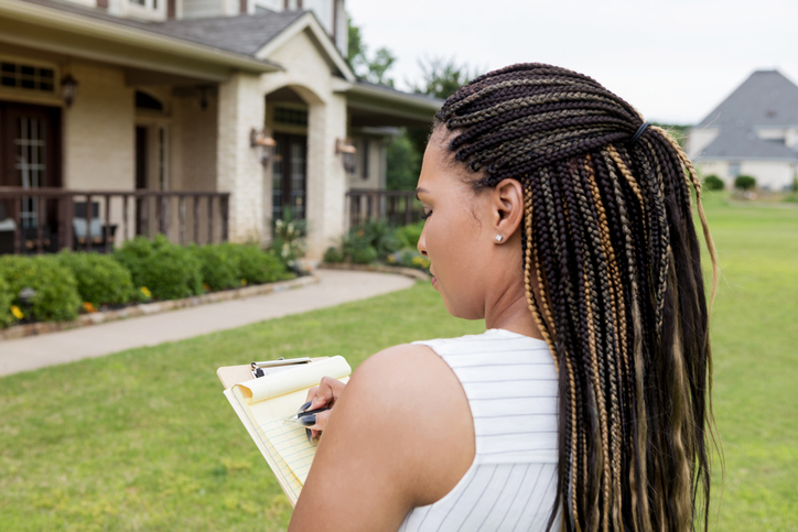 A female home inspector with dark skin and dark hair writes on a clipboard while she stands in front of a brown house.