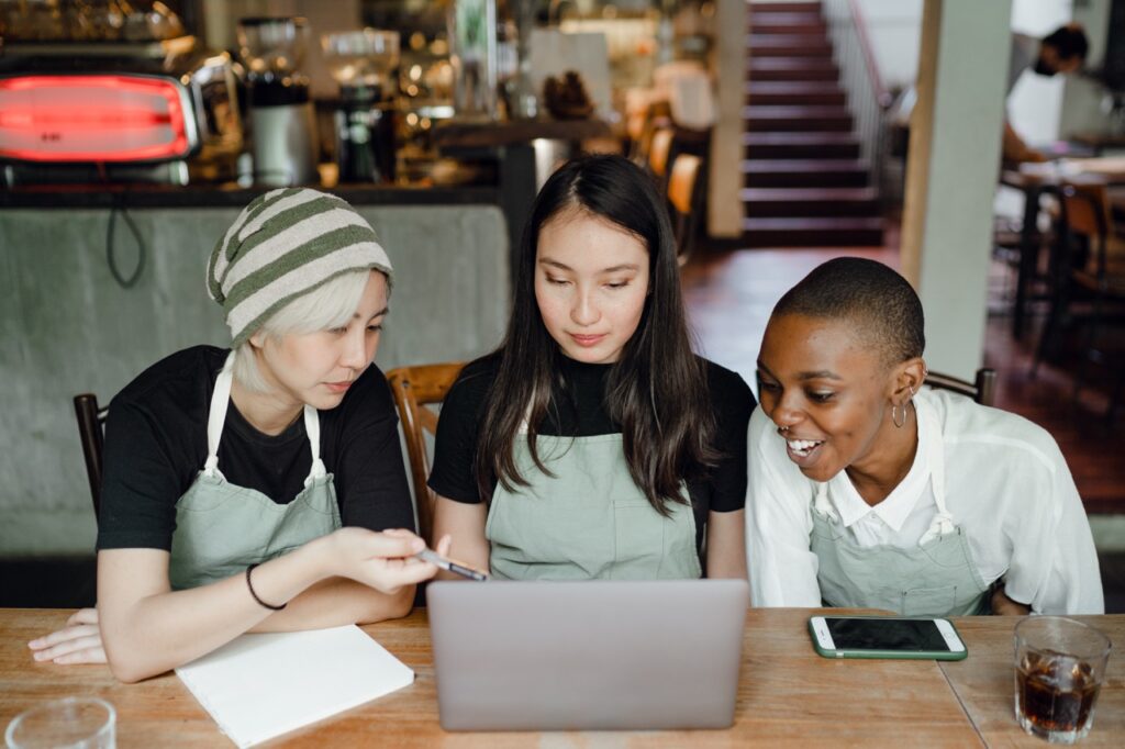 Three female-presenting business owners are sitting at a table looking at a laptop. Each person is wearing an apron and a coffee shop setting is in the background. The image implies that they are discussing business.