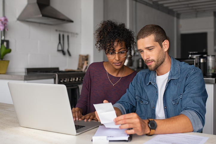 A young male and female couple are seated at a table and reviewing their finances in front of an open computer.