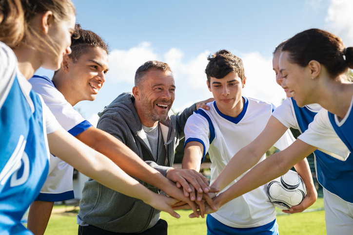 A youth soccer team and coach put their hands in the middle for a huddle.